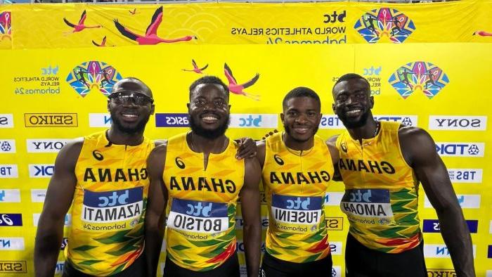 四名运动员在黄色背景前摆姿势, wearing their yellow Ghana jerseys and smiling with their arms around each other.